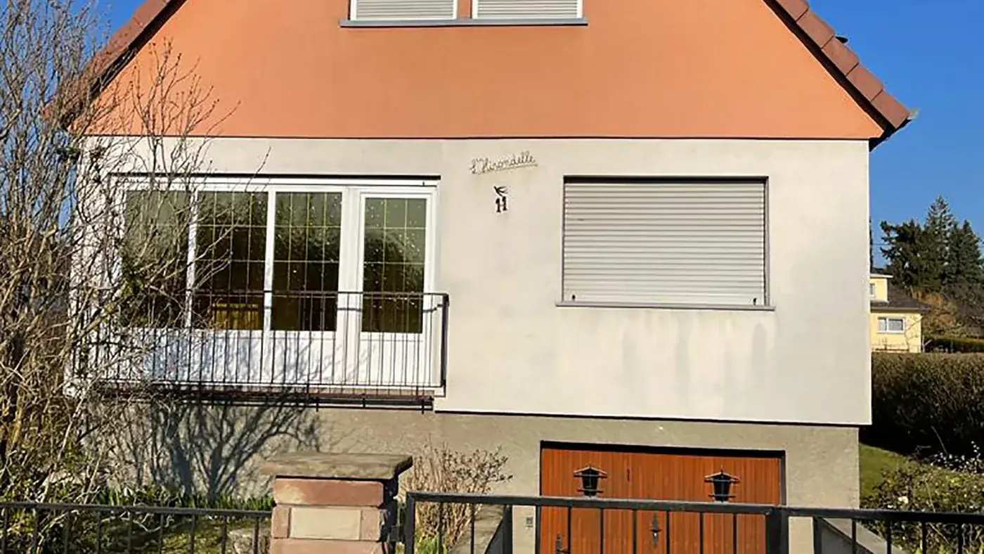 Maison alsacienne avant travaux : petites fenêtres blanches, garage en bois, volets roulants d'origine